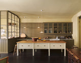 A simple Quaker cream and Folgate green Shaker kitchen features a cooker and sink run with Carrara marble worktop, glazed wall cupboards, and a bespoke prep table with a classic oak worktop. Lovingly adorned with milk pendant lights, artisan ceramics, antiques, and Helen stools, it creates a warm and inviting atmosphere.