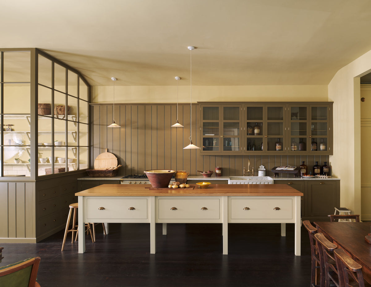 A simple Quaker cream and Folgate green Shaker kitchen features a cooker and sink run with Carrara marble worktop, glazed wall cupboards, and a bespoke prep table with a classic oak worktop. Lovingly adorned with milk pendant lights, artisan ceramics, antiques, and Helen stools, it creates a warm and inviting atmosphere.
