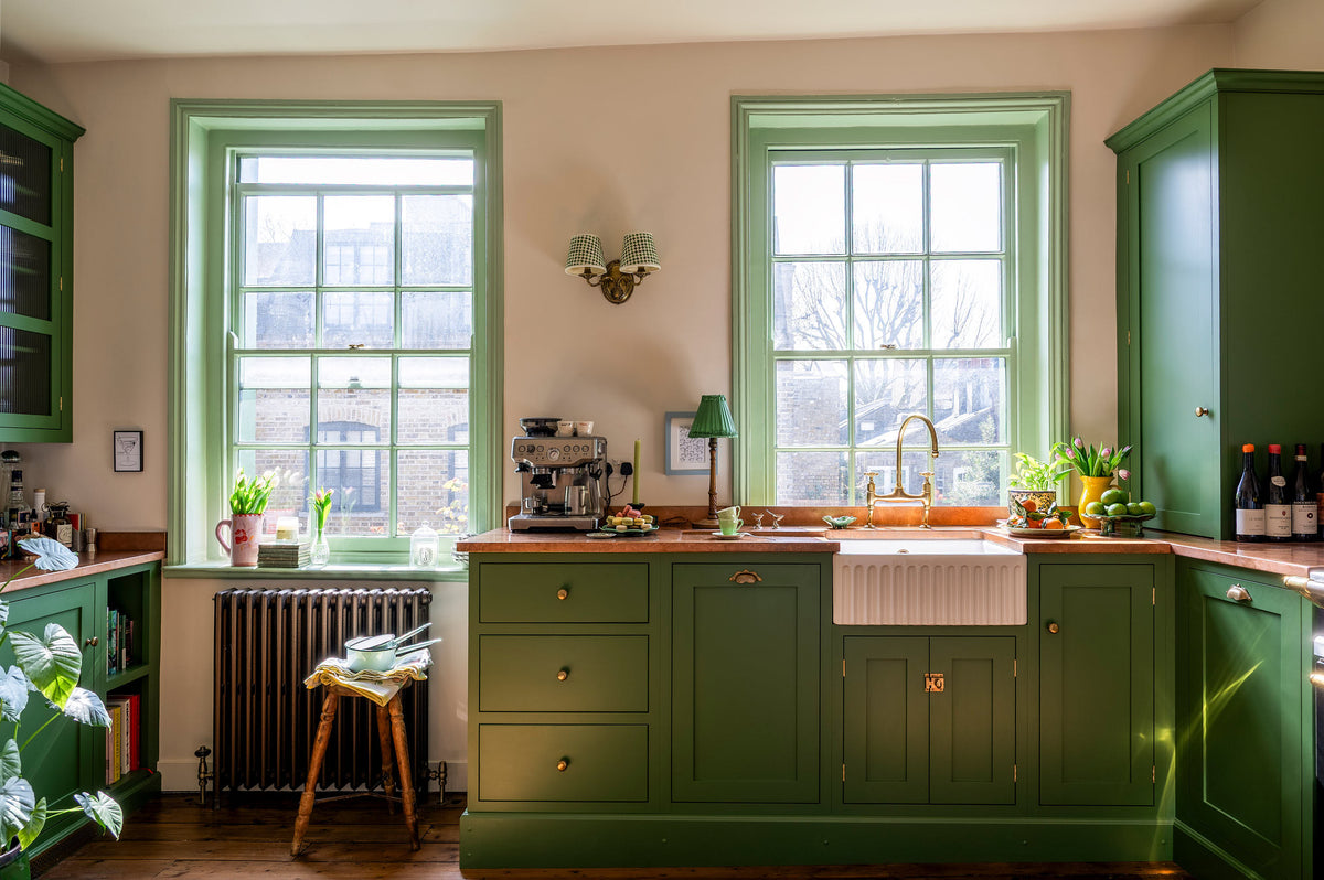 A bright and humble lime-green kitchen with an L-shaped cooker and sink run topped with aged copper worktops, featuring countertop and glazed cupboards, a single fluted Belfast sink with an aged brass Ionian tap, and colourful accents like tulips, a bowl of limes, macaroons, and assorted bottles of alcohol.