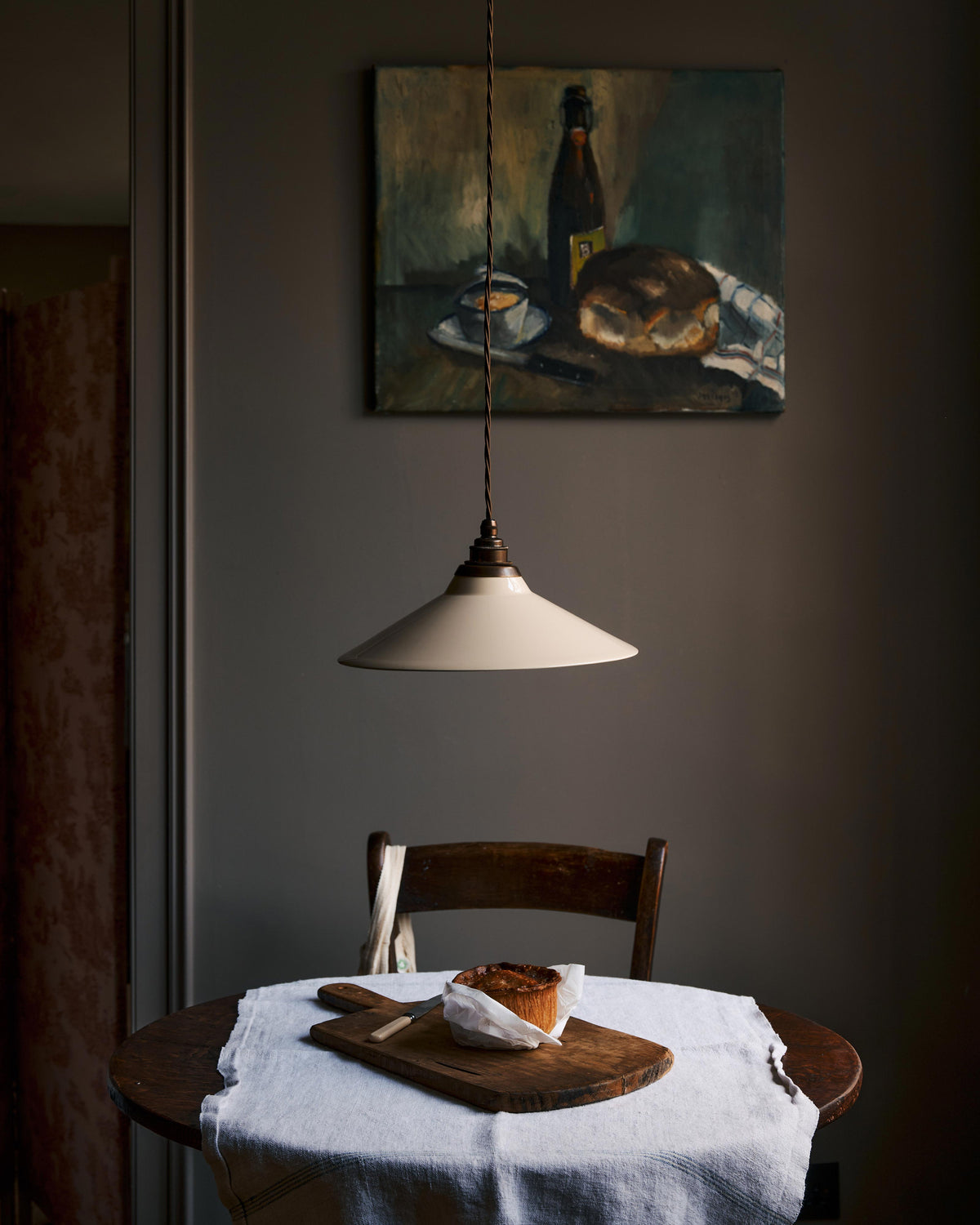 A straight-on view of a rise and fall light with a small cream glazed ceramic shade, aged brass fittings and brown twisted fabric flex cord hanging in a dark room over a small round wooden table with a linen tablecloth and fresh bread.