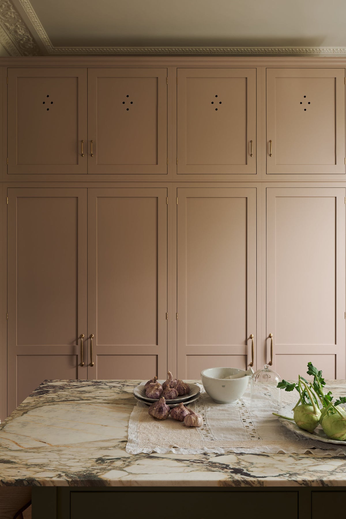A tall princelet pink cupboard pantry behind a marble top island with vegetables and ceramics.