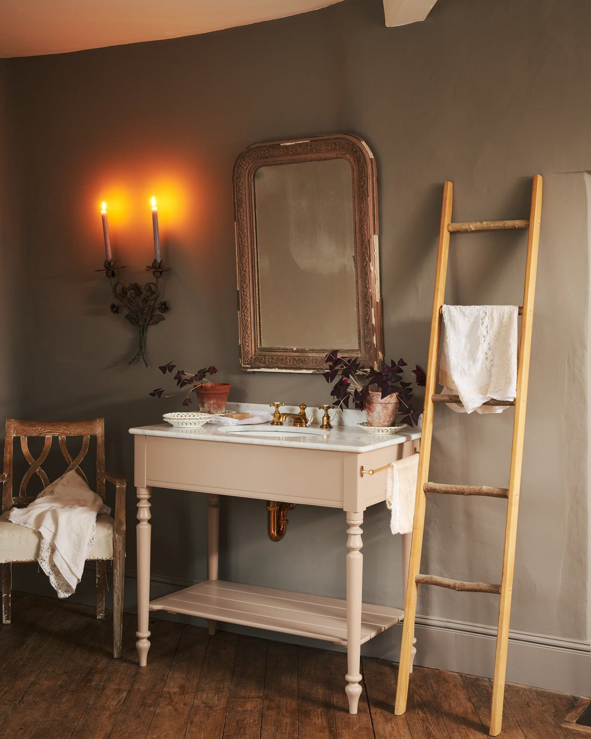 An oblique view towards a dusty-pink Victorian-inspired washstand with a brass mixer tap in a greyish-brown painted room. The shot is atmospheric and moody, with two lit candles casting a subtle glow and light from an adjacent window illuminating the brass accents. | Large Princelet Pink shown TEST