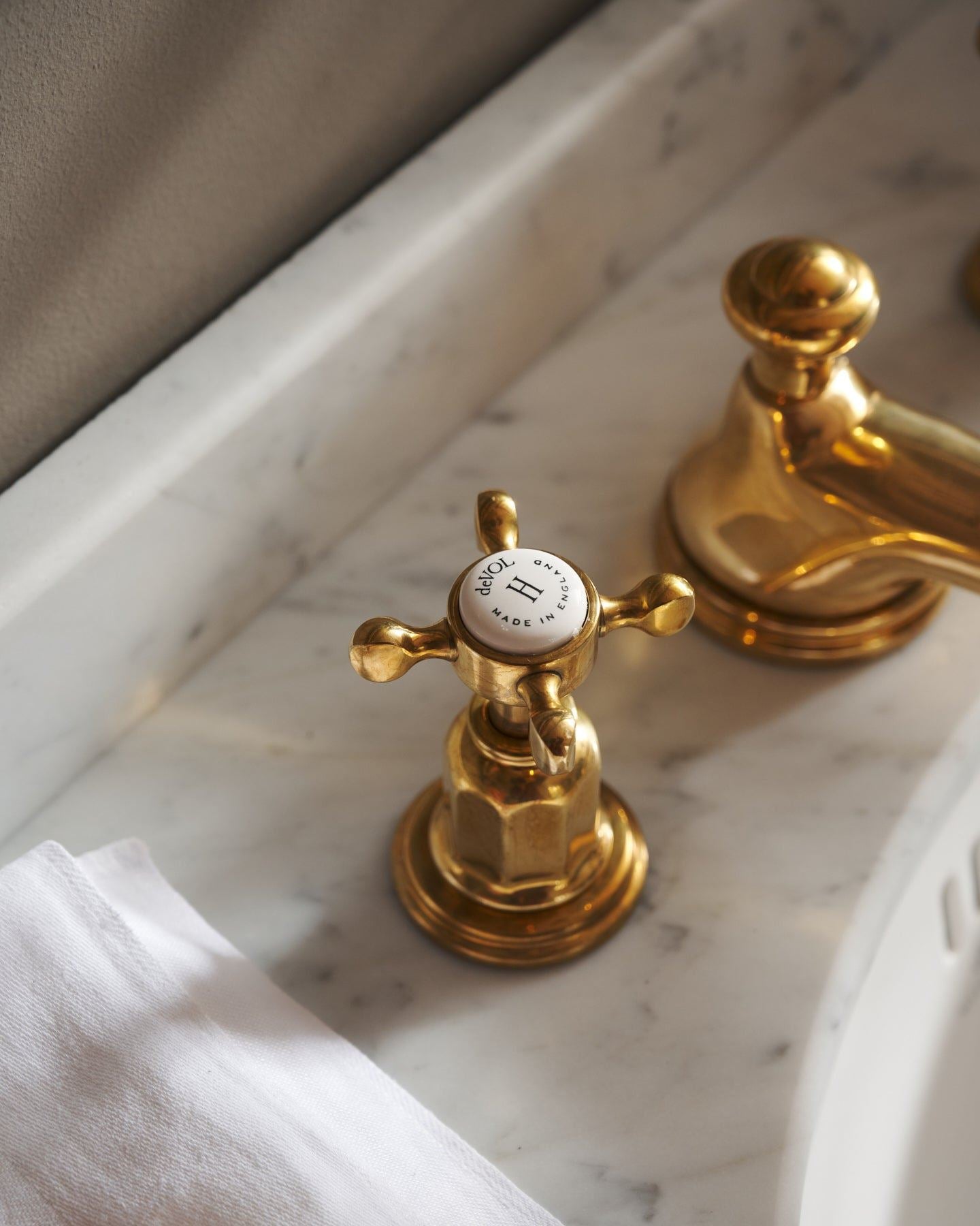 A close-up oblique view of the crosshead handle of an aged brass mixer tap for a bathroom basin sink. The letter 'H' is in the centre of the handle top, surrounded by the words 'deVOL, Made in England'.