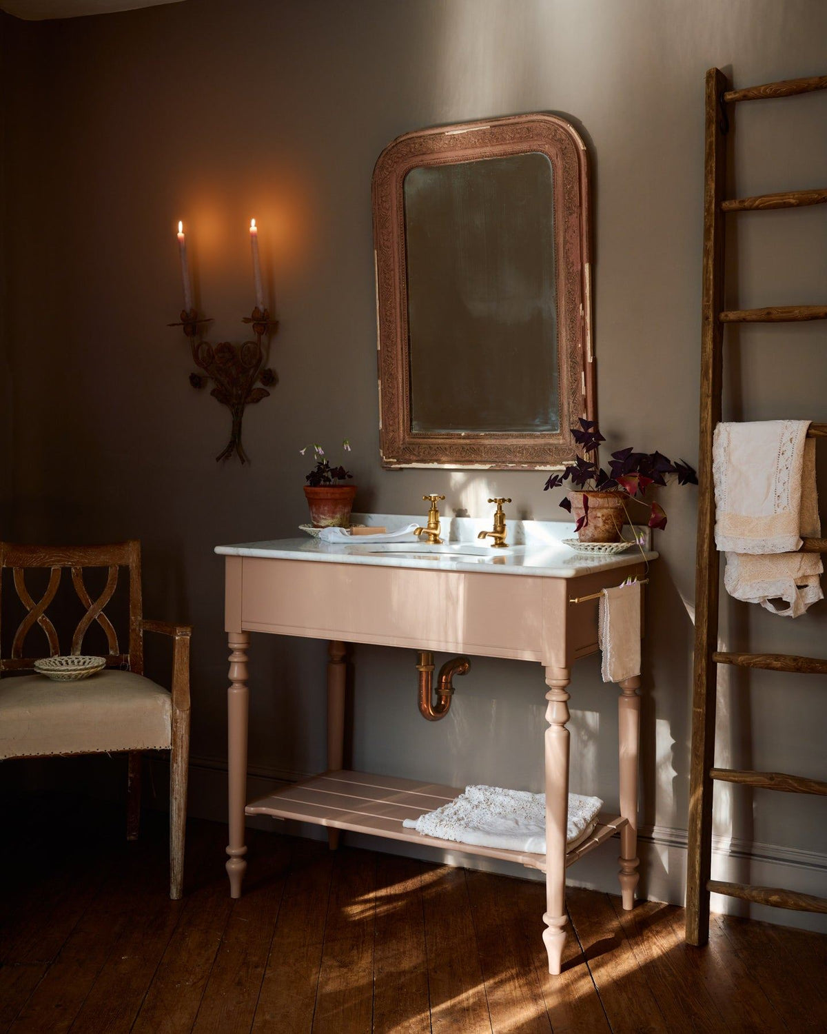 An oblique view towards a dusty-pink Victorian-inspired washstand with brass pillar taps in a greyish-brown painted room. The shot is atmospheric and moody, with two lit candles casting a subtle glow and light from an adjacent window illuminating the brass accents. | Large Princelet Pink shown TEST