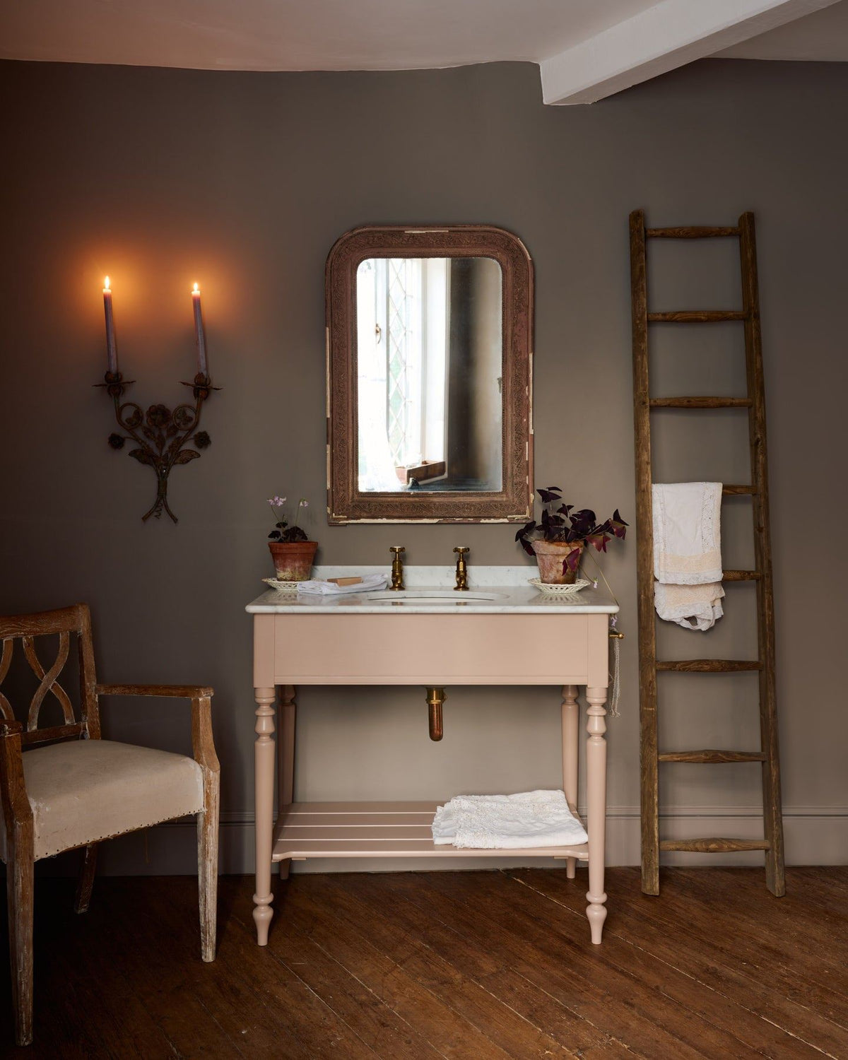 A straight-on view of a dusty-pink Victorian-inspired washstand with brass pillar taps in a greyish-brown painted room. The shot is atmospheric and moody, with two lit candles casting a subtle glow and light from an adjacent window illuminating the brass accents. TEST