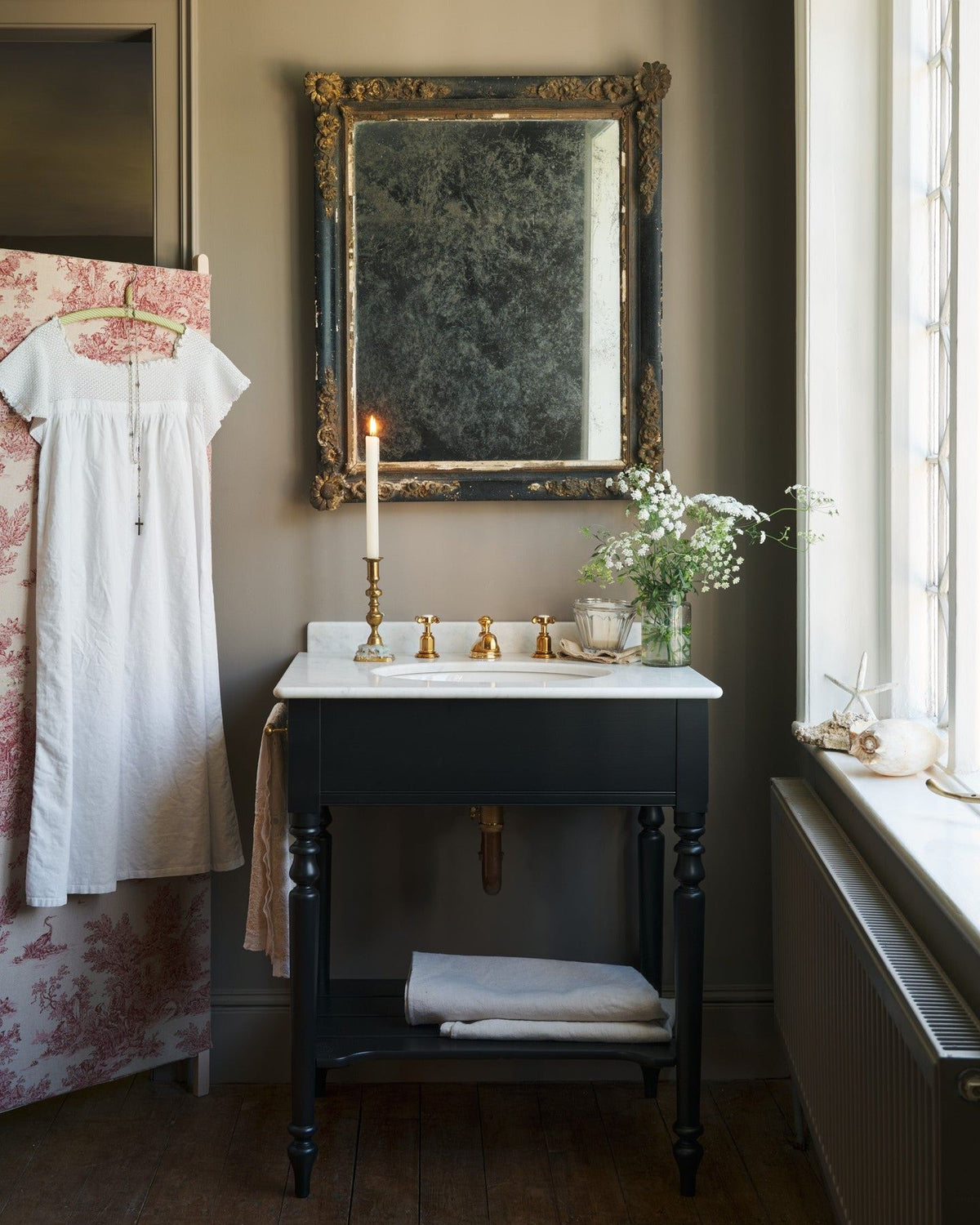 A straight-on view of a black Victorian-inspired washstand with a brass mixer tap in a greyish-brown painted room. A mottled antique mirror sits above the sink, an antique cotton dress hangs from a decorated partition and light streams through a nearby leaded window. | Medium Printer's Black shown TEST