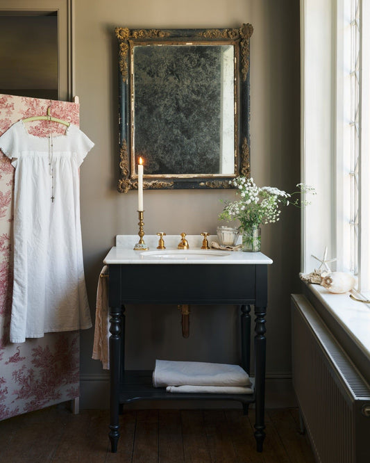 A straight-on view of a black Victorian-inspired washstand with a brass mixer tap in a greyish-brown painted room. A mottled antique mirror sits above the sink, an antique cotton dress hangs from a decorated partition and light streams through a nearby leaded window. | Medium Printer's Black shown