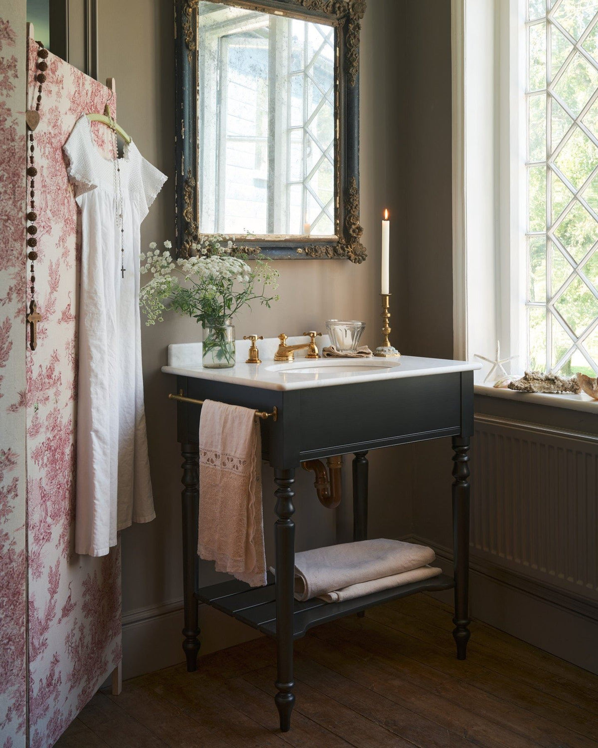 An oblique view of a black Victorian-inspired washstand with a brass mixer tap in a greyish-brown painted room. A mottled antique mirror sits above the sink, an antique cotton dress hangs from a decorated partition and light streams through a nearby leaded window. | Medium Printer's Black shown TEST
