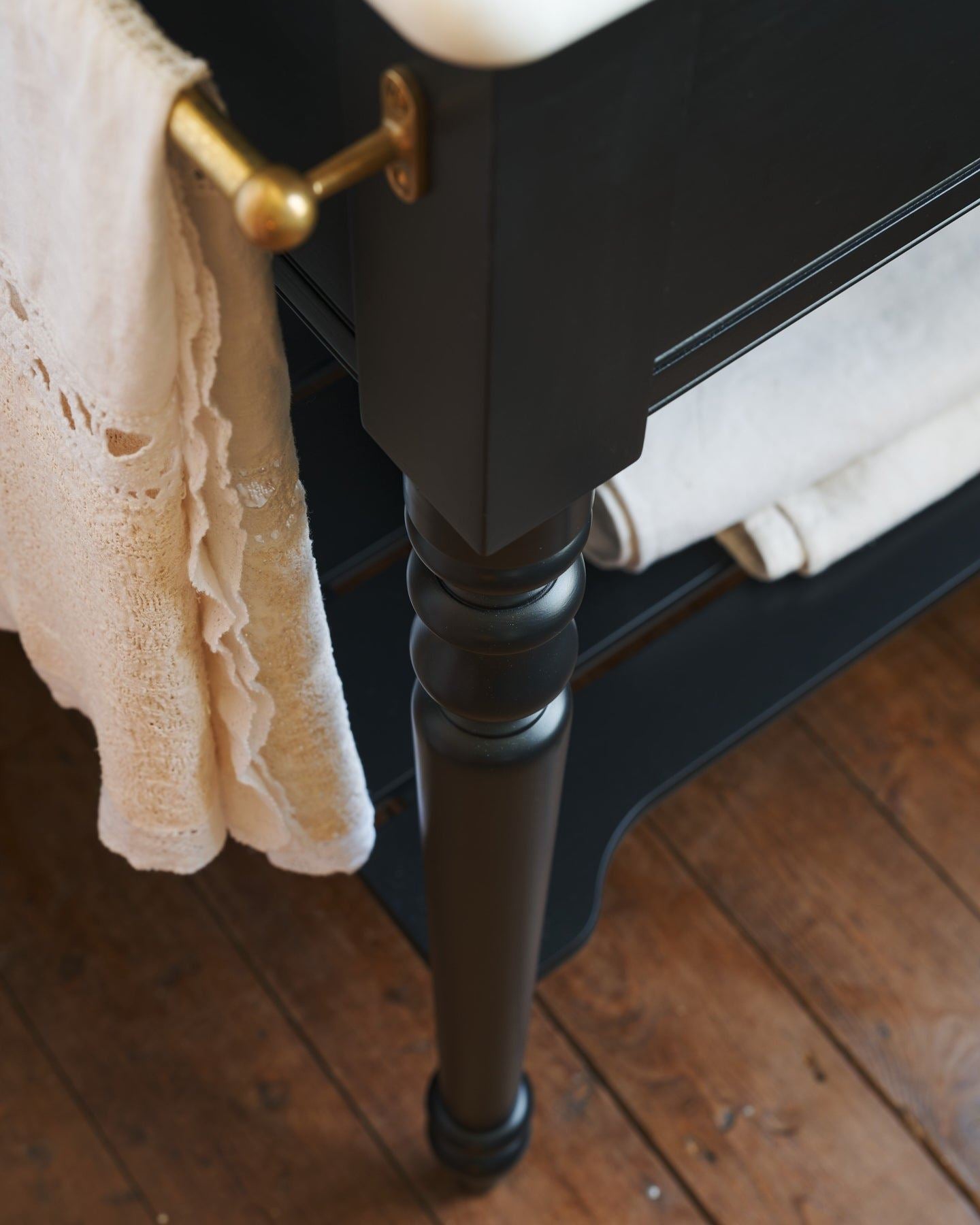 A detailed view of an aged brass towel rail attached to the left side of a Victorian-inspired washstand, a vintage lace hand towel hangs from the rail.