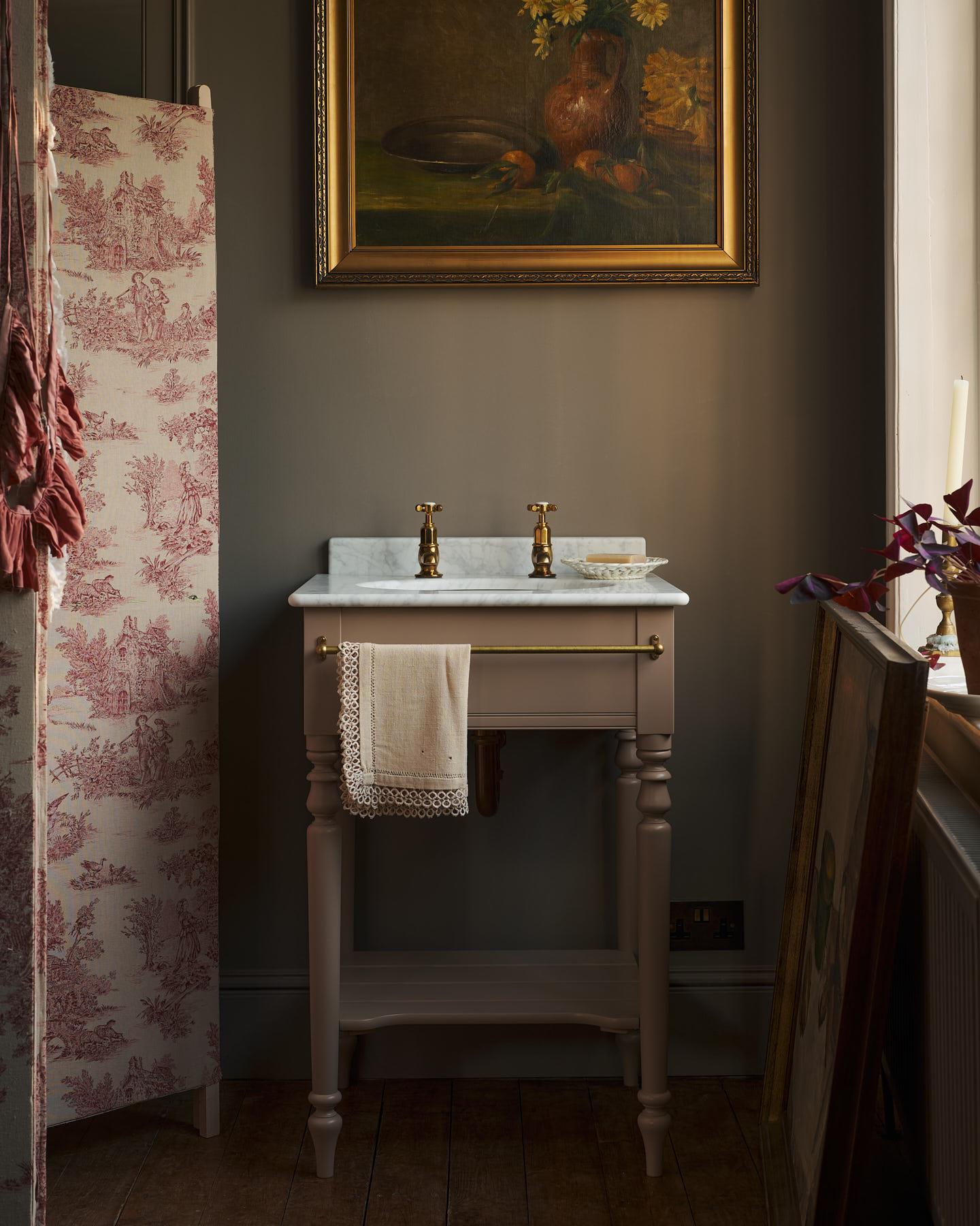 An oblique view towards a dusty-pink Victorian-inspired washstand with a brass mixer tap in a greyish-brown painted room. The shot is atmospheric and moody, with two lit candles casting a subtle glow and light from an adjacent window illuminating the brass accents.