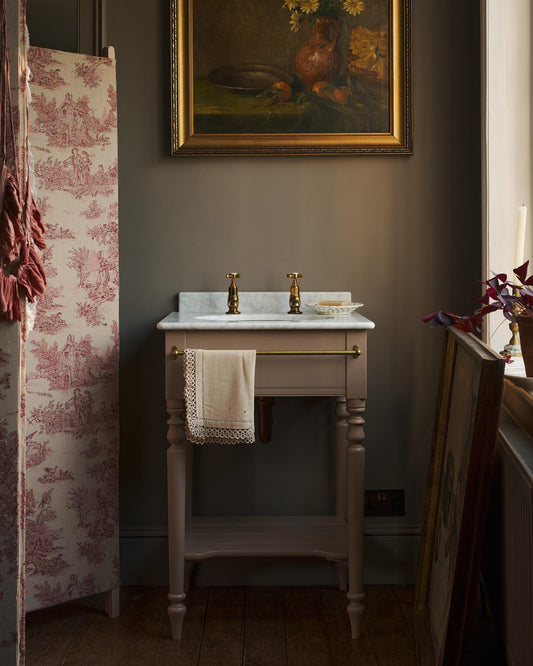 An oblique view towards a dusty-pink Victorian-inspired washstand with a brass mixer tap in a greyish-brown painted room. The shot is atmospheric and moody, with two lit candles casting a subtle glow and light from an adjacent window illuminating the brass accents. | Pillar Taps shown