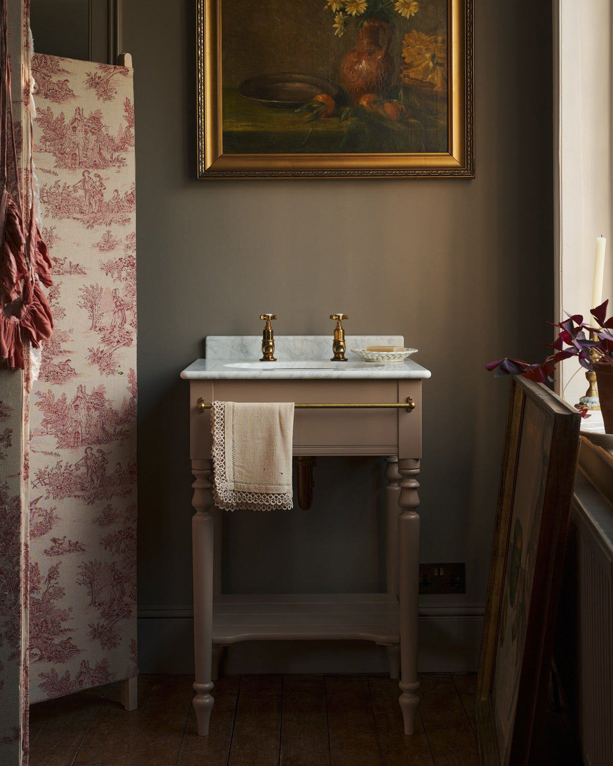 A straight-on view of a dusty-pink Victorian-inspired washstand with brass pillar taps in a greyish-brown painted room. The shot is atmospheric and moody, a vintage lace towel hangs from a brass rail attached to the front of the washstand. TEST