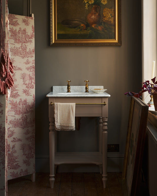 A straight-on view of a dusty-pink Victorian-inspired washstand with brass pillar taps in a greyish-brown painted room. The shot is atmospheric and moody, a vintage lace towel hangs from a brass rail attached to the front of the washstand.