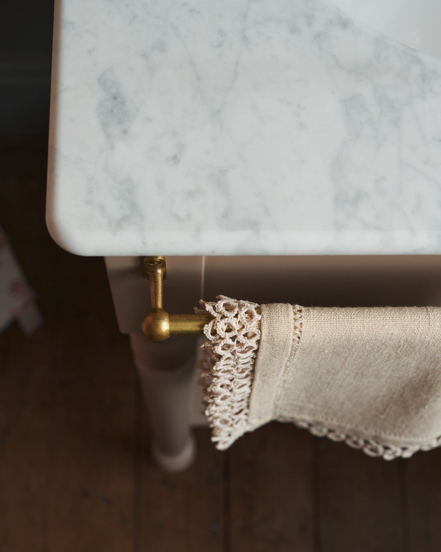 A detailed view of an aged brass towel rail attached to the front of a Victorian-inspired washstand, a vintage lace hand towel hangs from the rail.