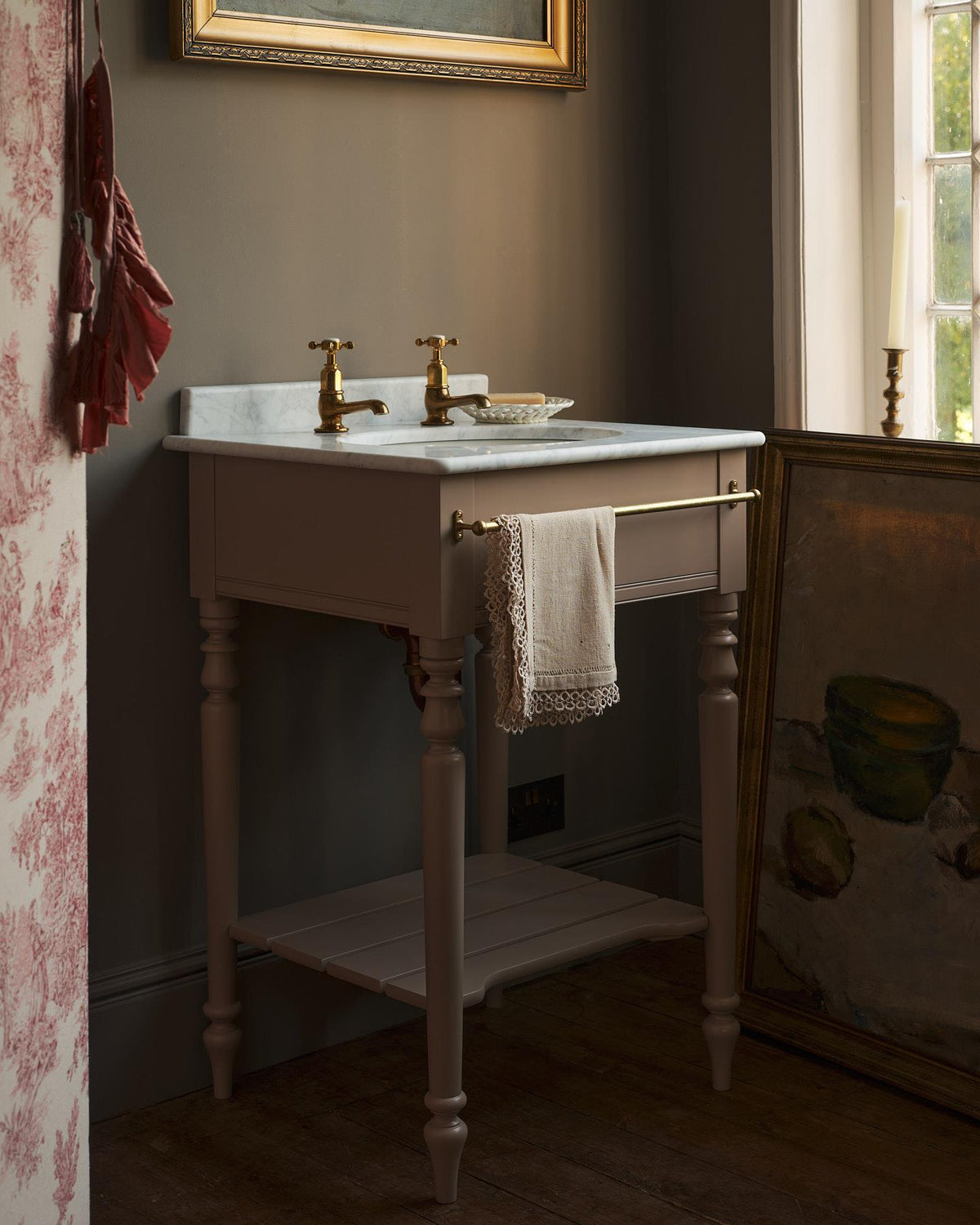 A straight-on view of a dusty-pink Victorian-inspired washstand with a brass mixer tap in a greyish-brown painted room. The shot is atmospheric and moody, with two lit candles casting a subtle glow and light from an adjacent window illuminating the brass accents. | Pillar Taps shown TEST