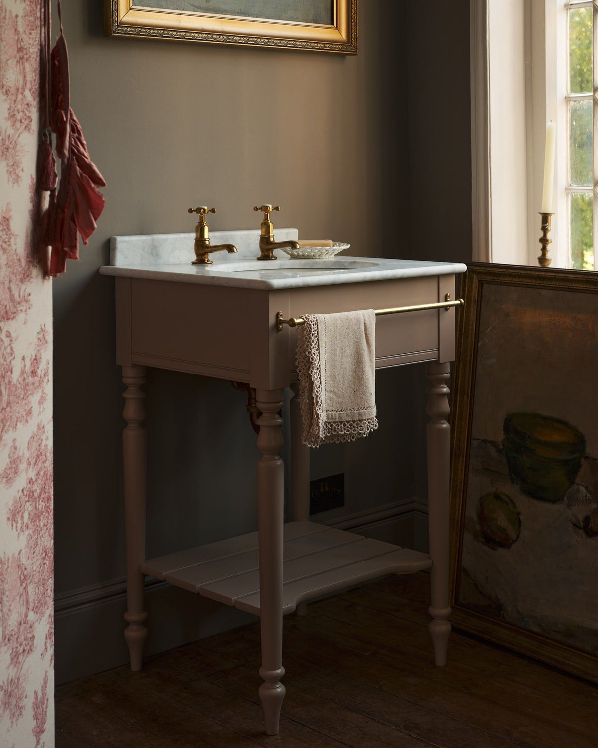 An oblique view of a dusty-pink Victorian-inspired washstand with brass pillar taps in a greyish-brown painted room. The shot is atmospheric and moody, a vintage lace towel hangs from a brass rail attached to the front of the washstand. TEST