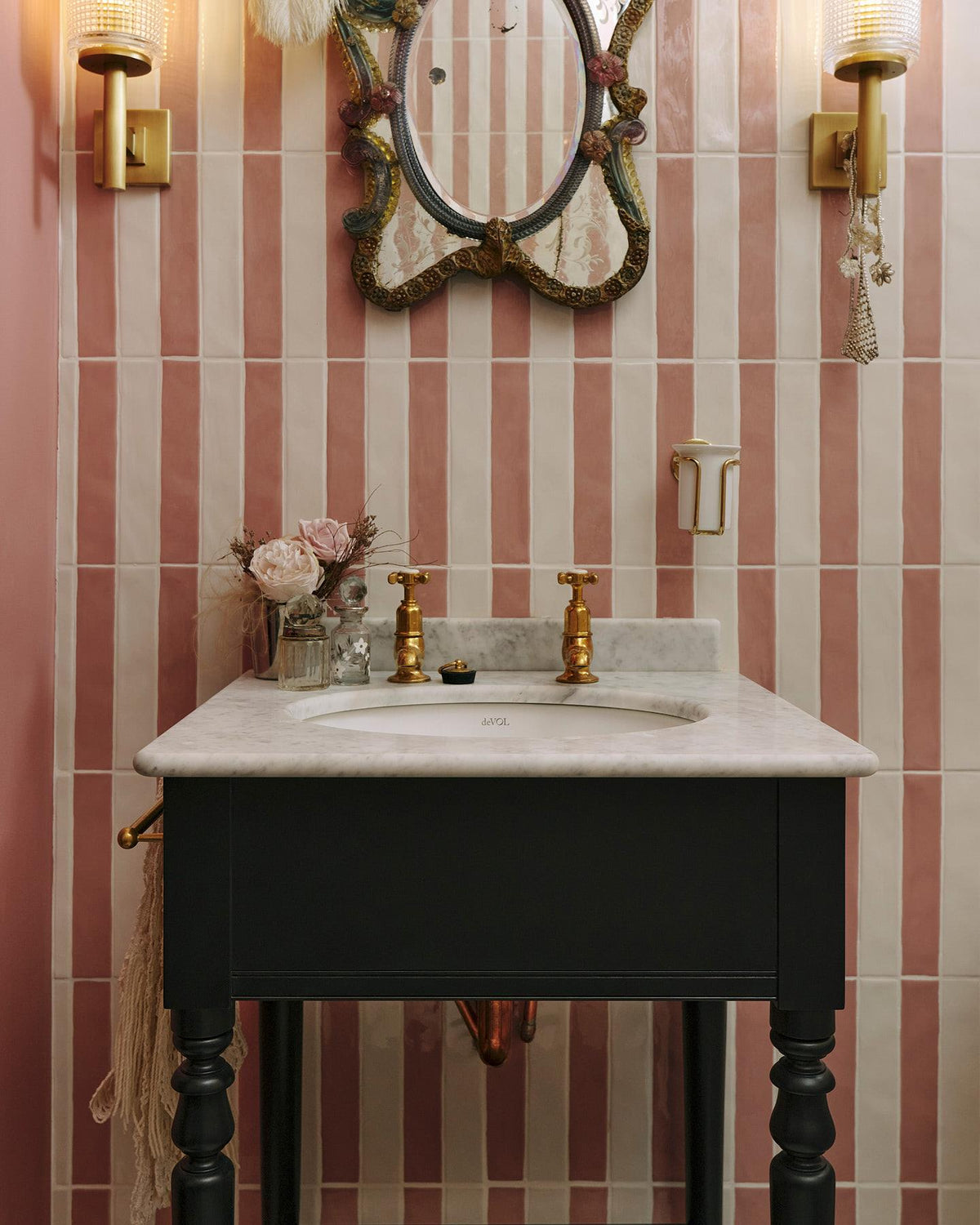 A straight-on view of a black Victorian-inspired washstand with a brass pillar tap against a pink and white splashback tiles. A decorative antique mirror sits above the sink, along with deco-style scones, and a brass cup holder on the wall. TEST