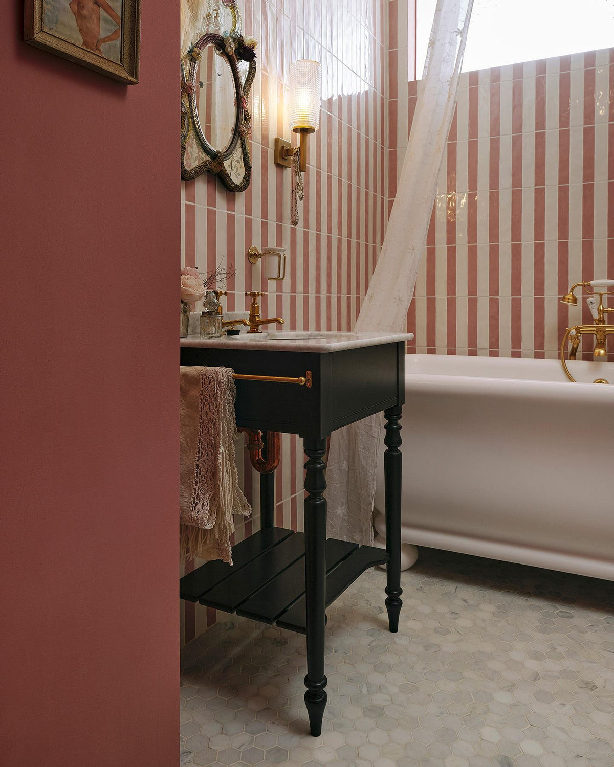 An oblique view of a porcelain bath and a black Victorian-inspired washstand with a brass pillar tap against a pink and white splashback tiles. A decorative antique mirror sits above the sink, along with deco-style scones, and a brass cup holder on the wall. TEST