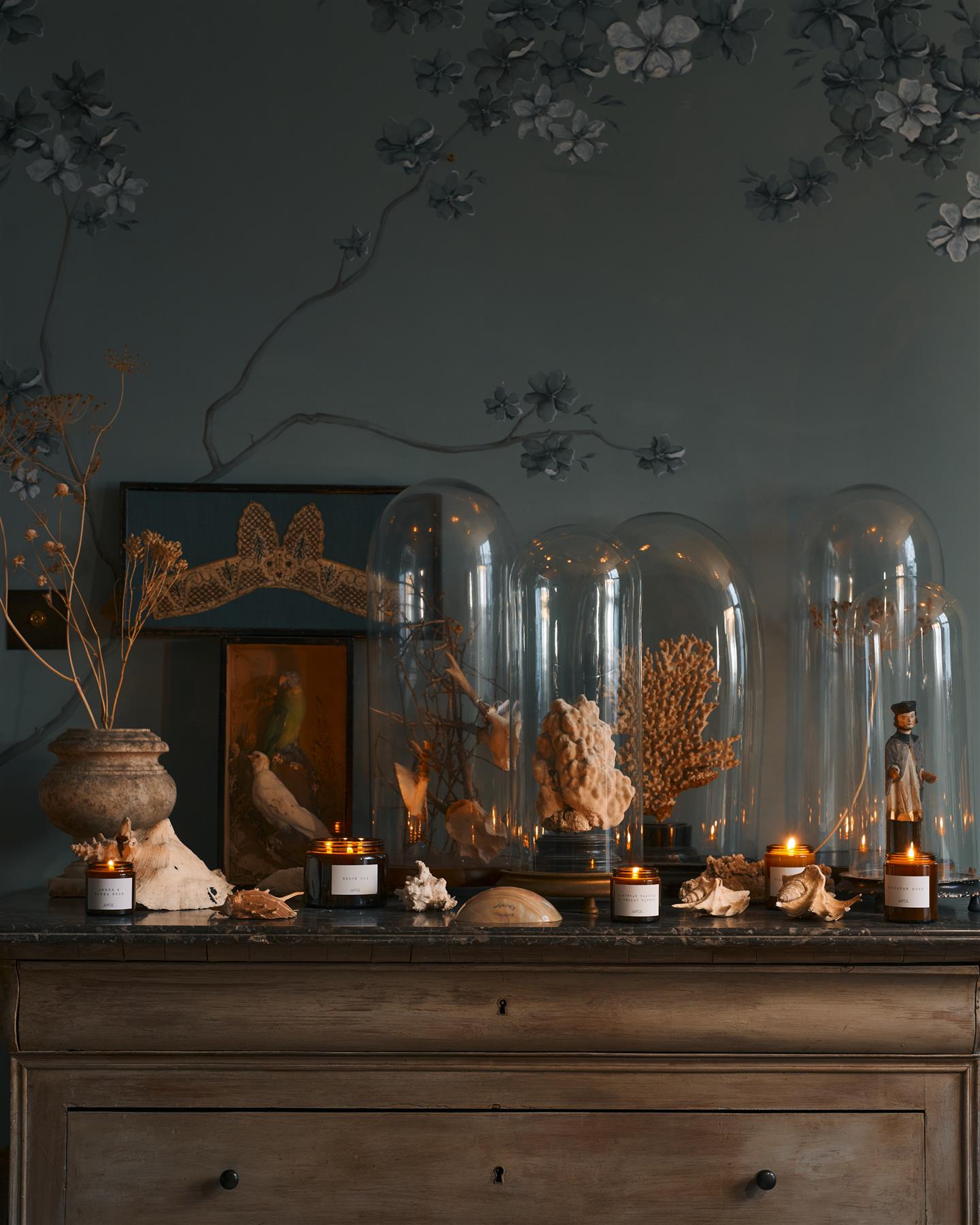 A medium scented candle in a brown glass jar with a black lid, in a vintage cupboard alongside other scented candles.