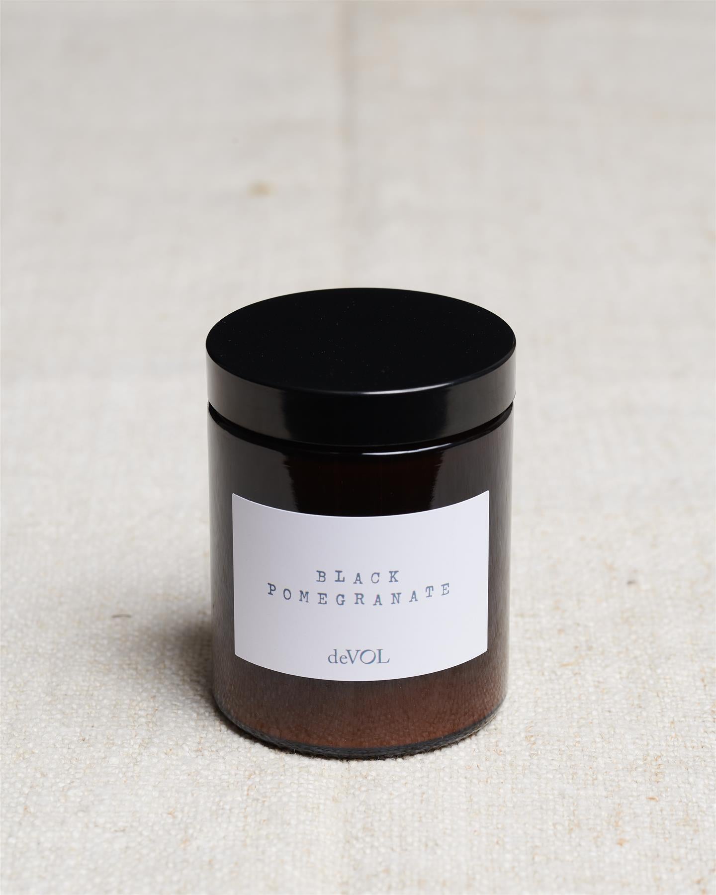 A medium scented candle in a brown glass jar with a black lid, the label reads 'Amber & Tonka Bean, deVOL'.