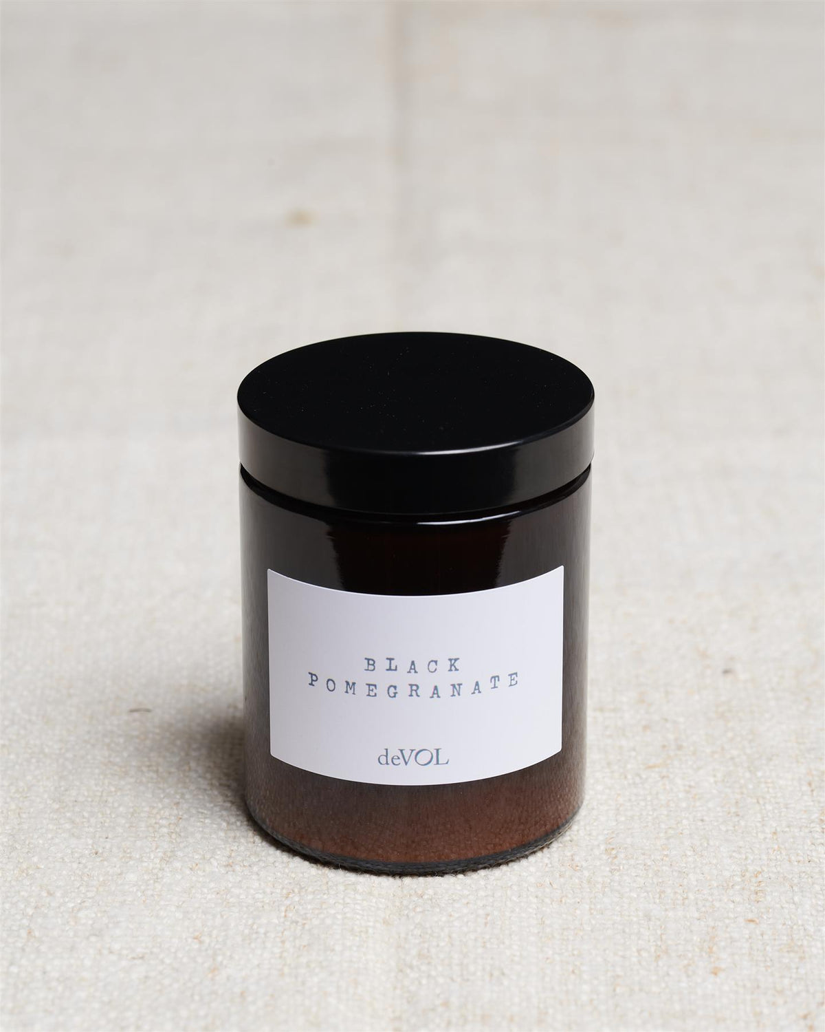 A medium scented candle in a brown glass jar with a black lid, the label reads 'Amber & Tonka Bean, deVOL'. TEST