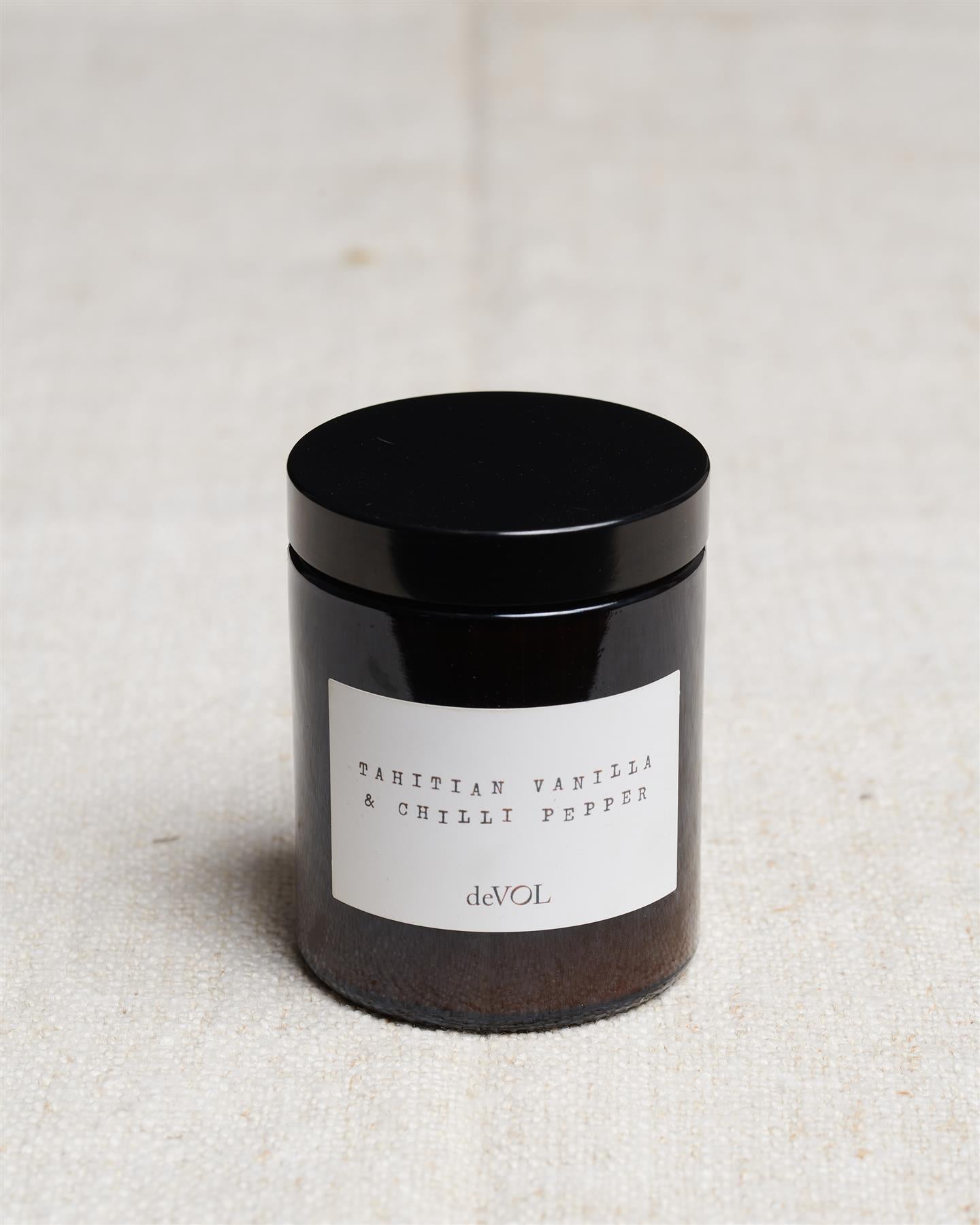 A medium scented candle in a brown glass jar with a black lid, the label reads 'Amber & Tonka Bean, deVOL'.