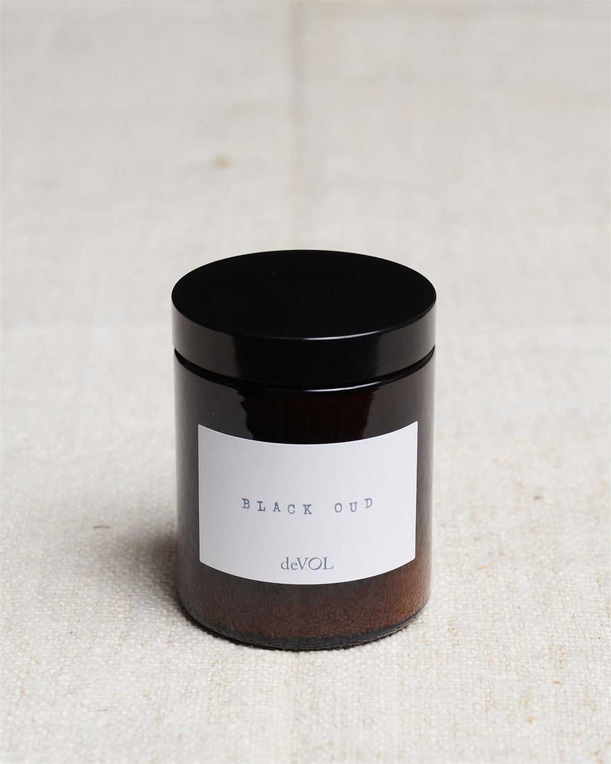 A medium scented candle in a brown glass jar with a black lid, the label reads 'Amber & Tonka Bean, deVOL'. TEST