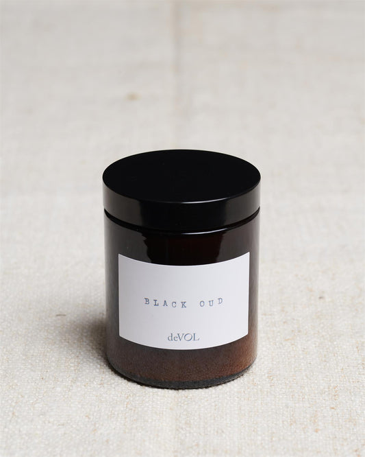 A medium scented candle in a brown glass jar with a black lid, the label reads 'Amber & Tonka Bean, deVOL'.