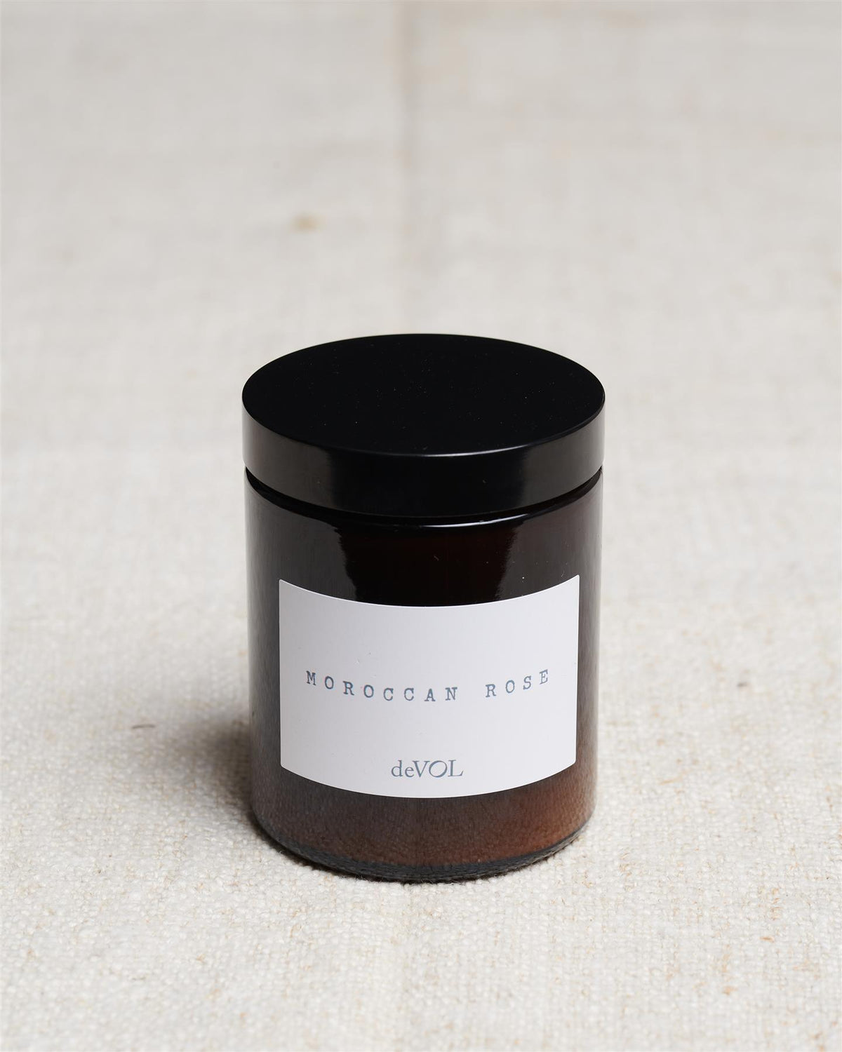 A medium scented candle in a brown glass jar with a black lid, the label reads 'Amber & Tonka Bean, deVOL'. TEST