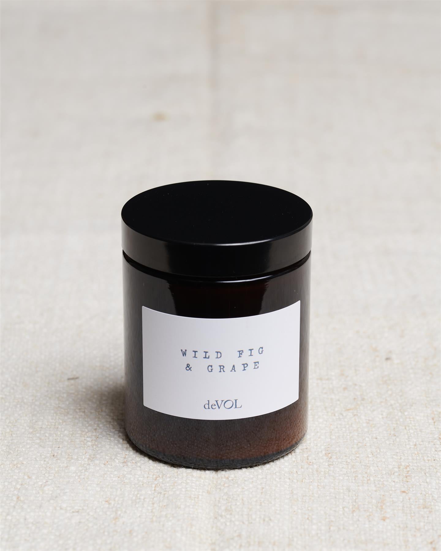 A medium scented candle in a brown glass jar with a black lid, the label reads 'Amber & Tonka Bean, deVOL'.