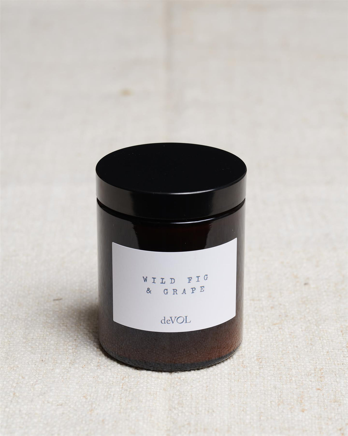 A medium scented candle in a brown glass jar with a black lid, the label reads 'Amber & Tonka Bean, deVOL'. TEST