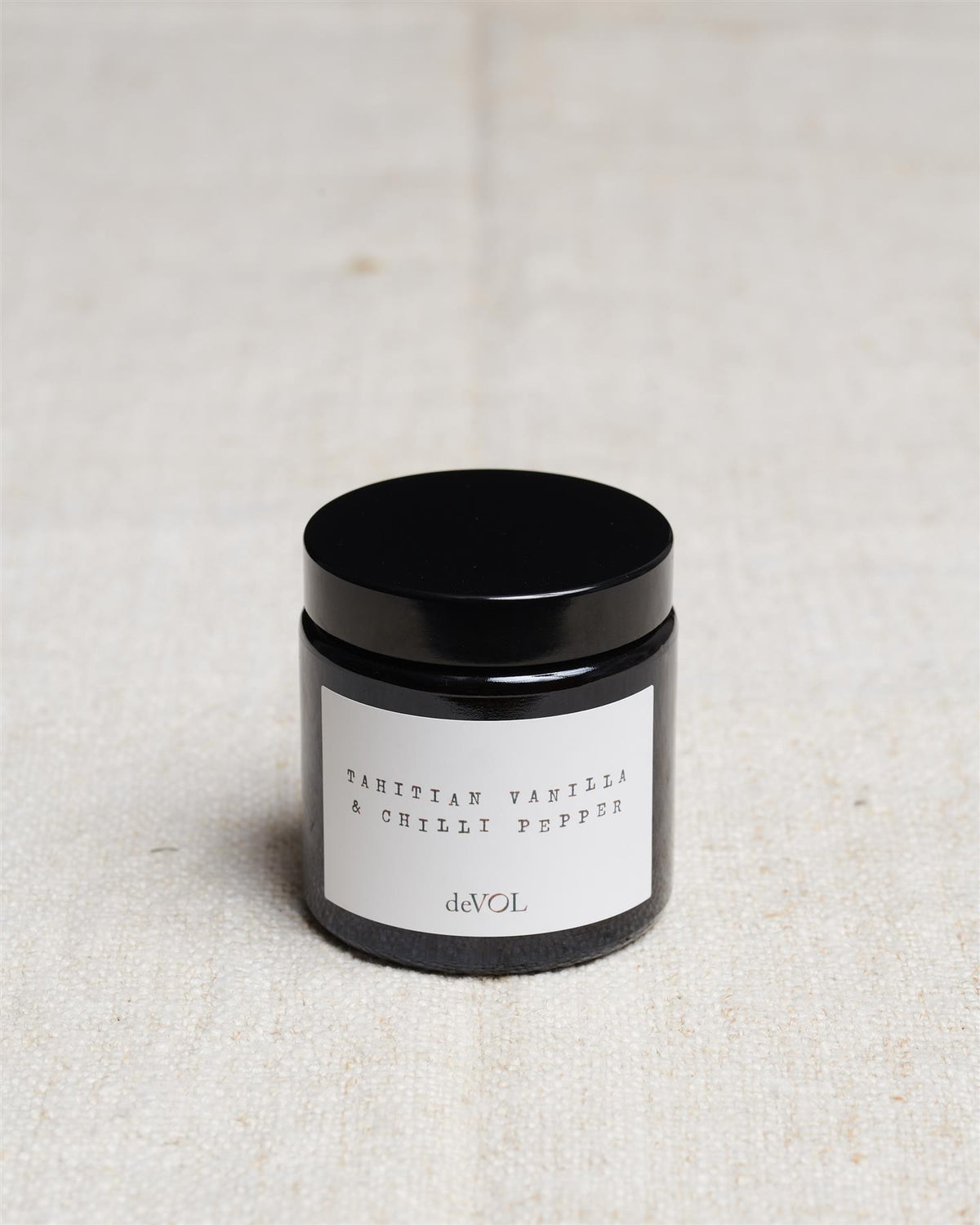 A small scented candle in a brown glass jar with a black lid, the label reads 'Amber & Tonka Bean, deVOL'. TEST