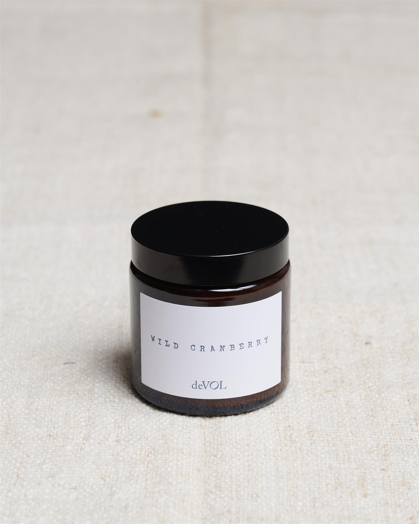 A small scented candle in a brown glass jar with a black lid, the label reads 'Amber & Tonka Bean, deVOL'.
