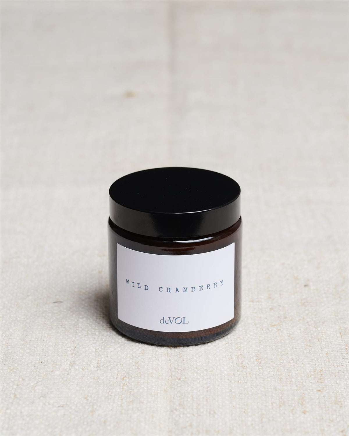 A small scented candle in a brown glass jar with a black lid, the label reads 'Amber & Tonka Bean, deVOL'. TEST