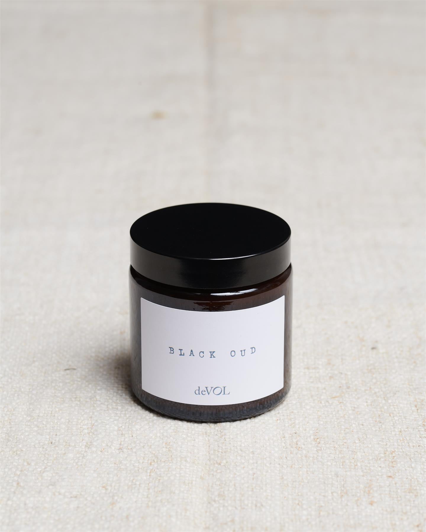 A small scented candle in a brown glass jar with a black lid, the label reads 'Amber & Tonka Bean, deVOL'.