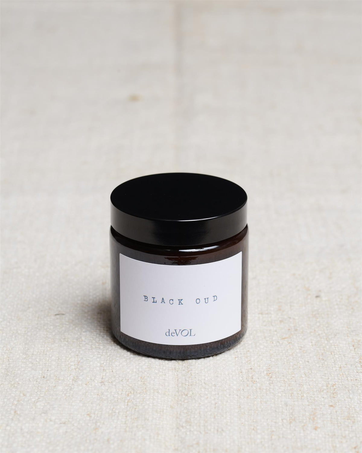 A small scented candle in a brown glass jar with a black lid, the label reads 'Amber & Tonka Bean, deVOL'. TEST