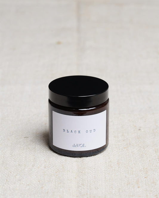 A small scented candle in a brown glass jar with a black lid, the label reads 'Amber & Tonka Bean, deVOL'.
