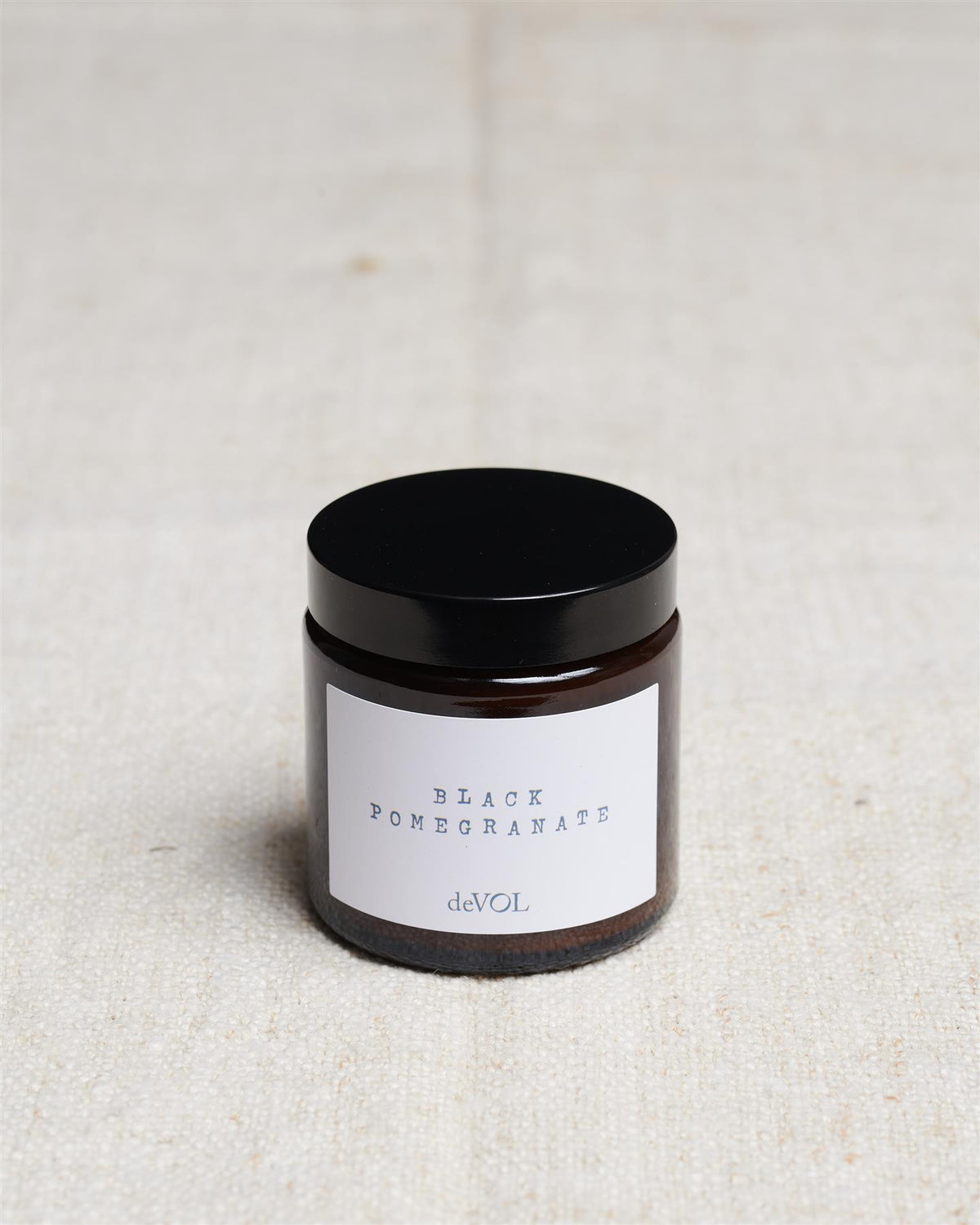 A small scented candle in a brown glass jar with a black lid, the label reads 'Amber & Tonka Bean, deVOL'. TEST