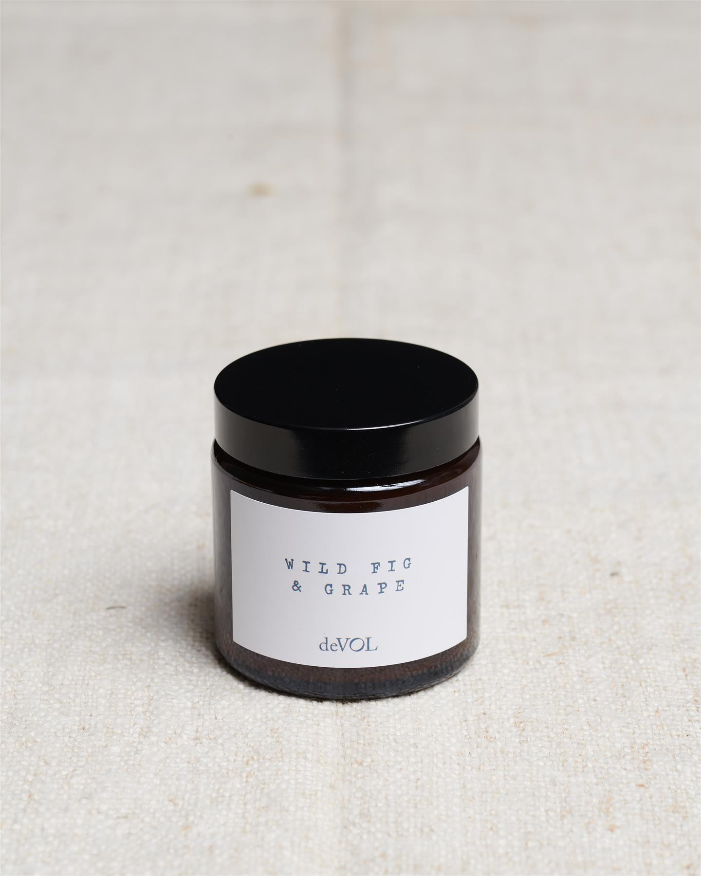 A small scented candle in a brown glass jar with a black lid, the label reads 'Amber & Tonka Bean, deVOL'.