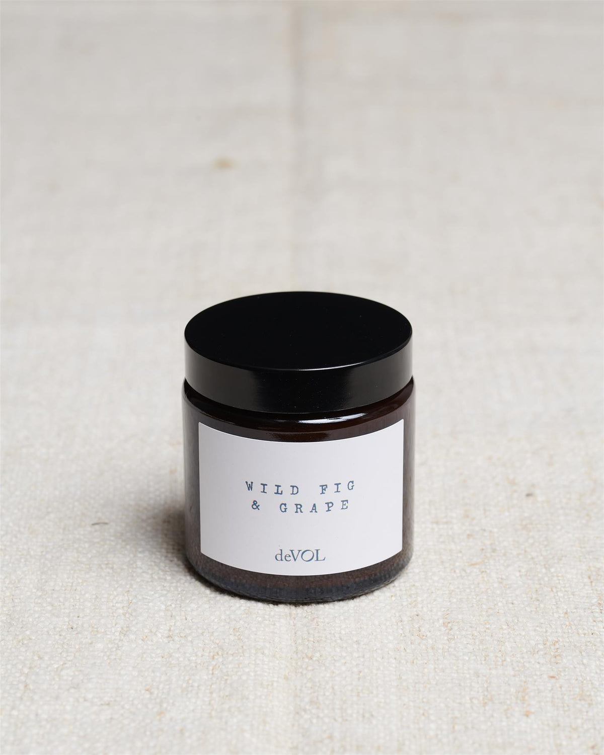 A small scented candle in a brown glass jar with a black lid, the label reads 'Amber & Tonka Bean, deVOL'. TEST
