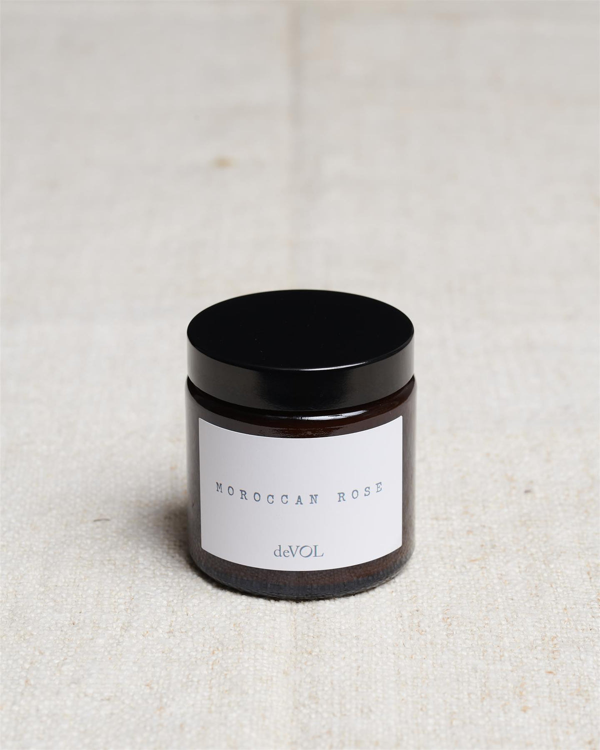 A small scented candle in a brown glass jar with a black lid, the label reads 'Amber & Tonka Bean, deVOL'. TEST