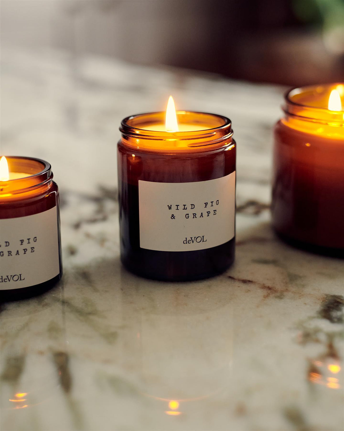 A straight-on view of a collection of three lit scented candles in brown glass jars on a wooden stool top; small, medium and large. TEST