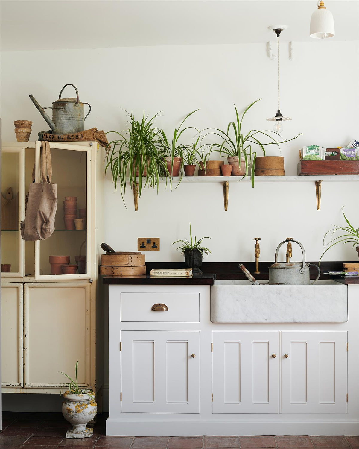 A straight-on view of a classic 13 amp twin plug socket in a mottled aged brass finish in a Mediterannean-inspired kitchen with a white painted Georgian-style sink cabinet, dark hardwood worktop, Carrara marble sink and a shelf full of lush spider plants.|Aged Brass shown TEST