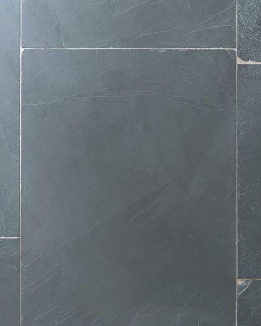 A close-up overhead view of a grey tumbled slate floor, with two loose tiles stacked in one corner.