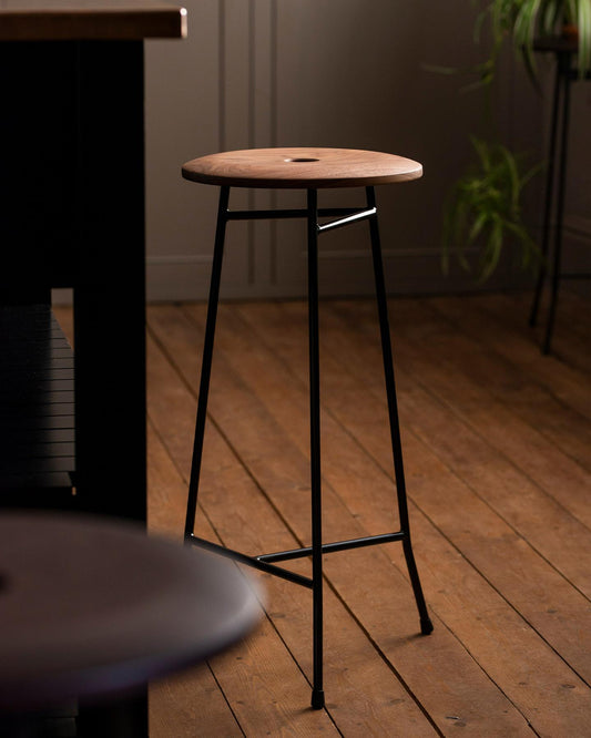 A mid-century-style three-legged bar stool with a thin dark metal frame and a round walnut seat top with a central hole and a natural oiled finish.