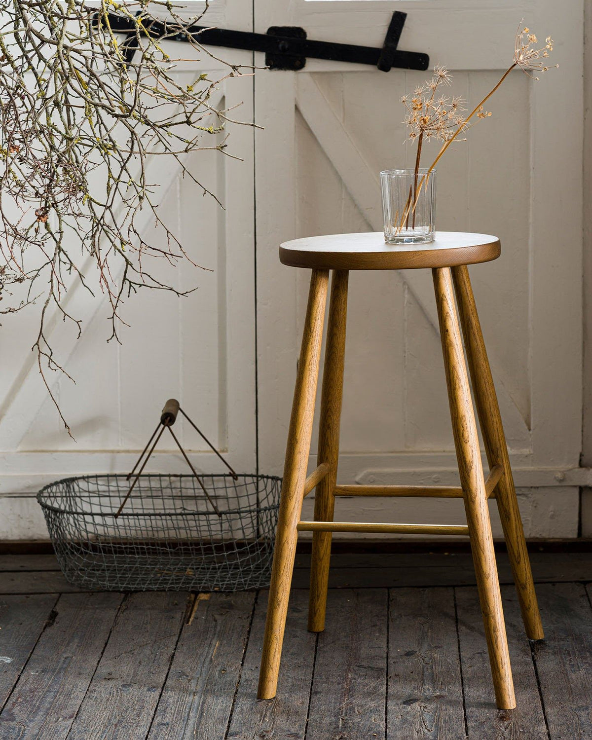 A straight-on view of a simple oak bar stool with an oval-shaped seat and a brass footrail with dried flowers in a glass on top. TEST