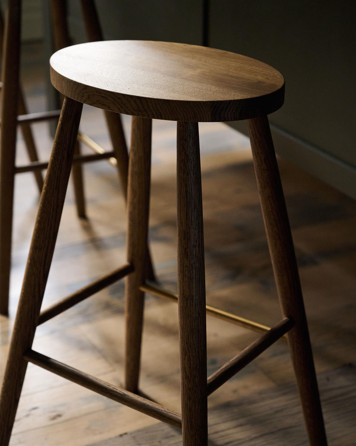 An oblique view of a simple oak bar stool with an oval-shaped seat and a brass footrail. TEST