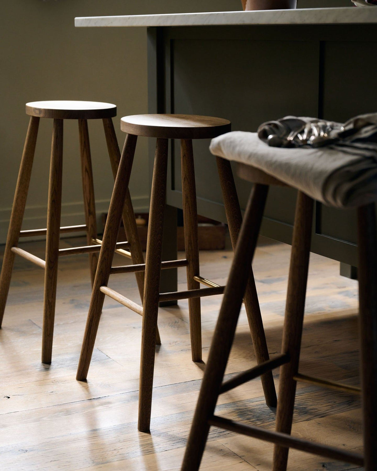 An oblique view of threee simple oak bar stools with oval-shaped seats in front of an island breakfast bar. TEST