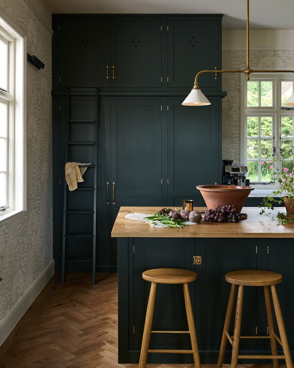 A straight-on view of a dark green painted Shaker-style kitchen with an island breakfast bar and two simple oak bar stools with oval-shaped seats and brass footrails. TEST