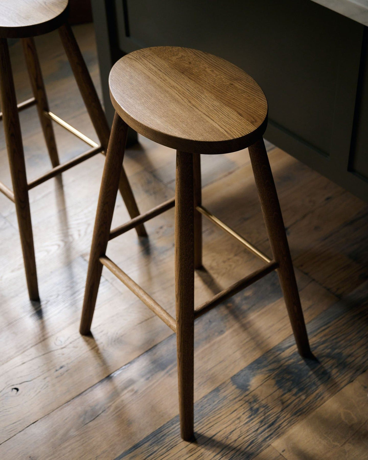 An oblique view of three simple oak bar stools with oval-shaped seats in front of a dark green painted Shaker-style island breakfast bar. TEST